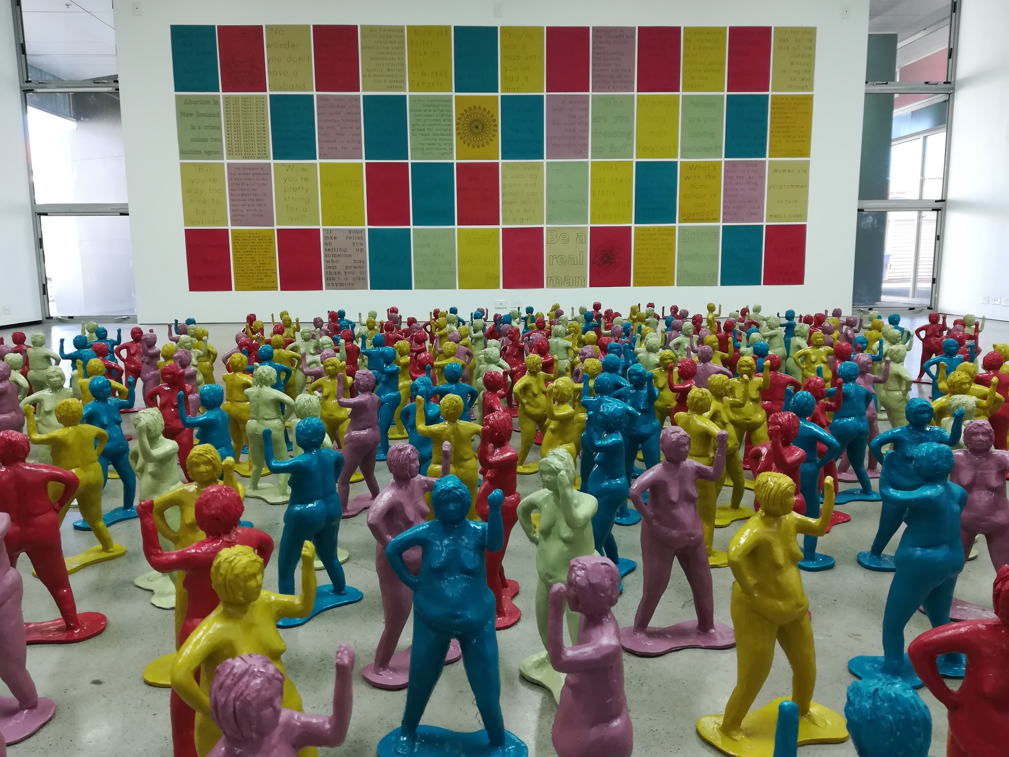 Photo of art installation. 300 small ceramic bertha figures in front of a wall of feminist posters