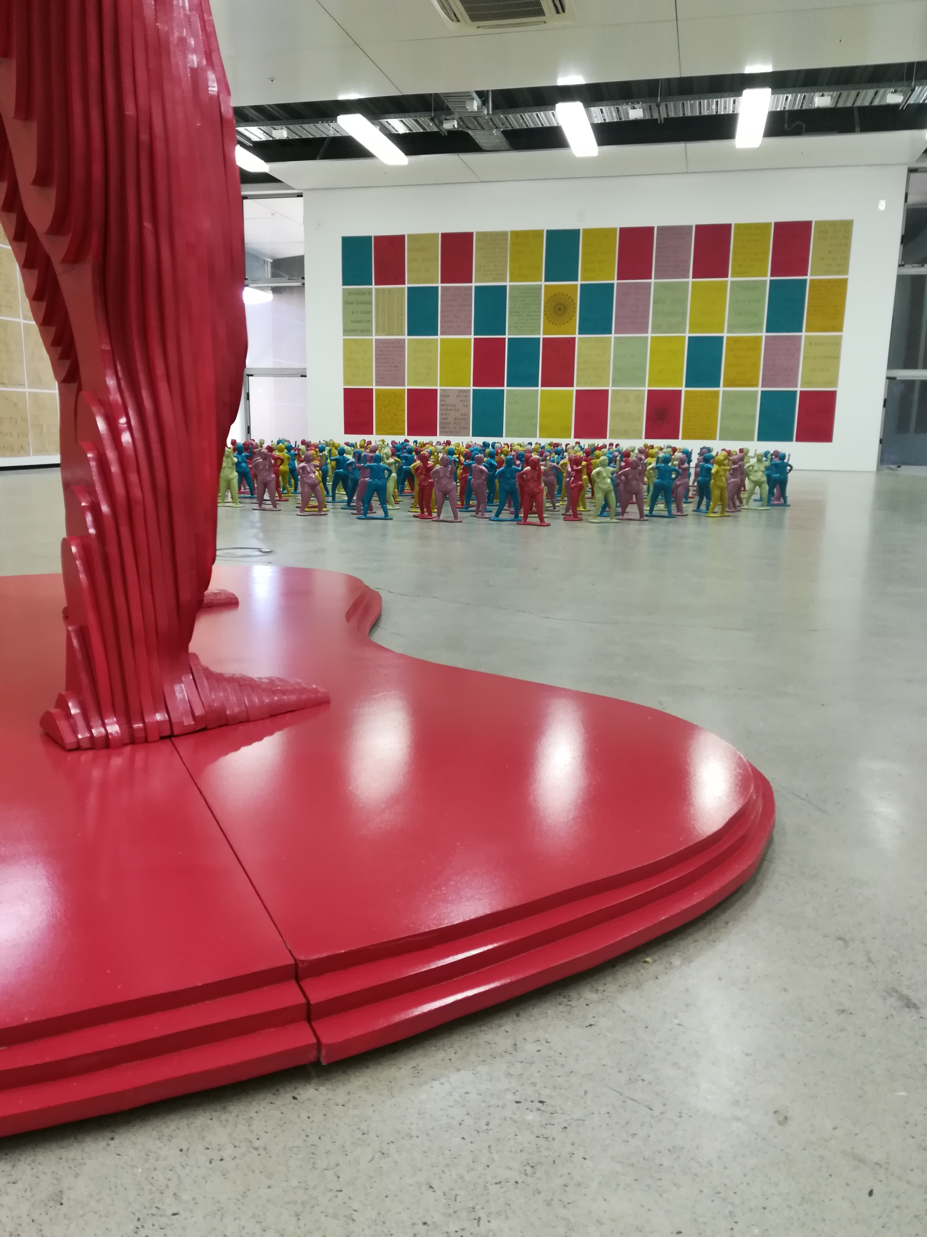 Photo of exhibition installation. Background is 300 ceramic bertha figures with posters on the wall. Foreground is the legs of the 3m tall wooden Bertha.